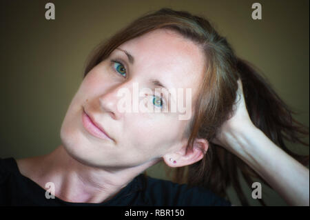 A young woman her head tilted sideways resting in her hand.  Her long hair in a ponytail her freckled blue green eyes looking into camera Stock Photo