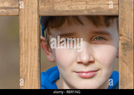 A young boy with beautiful green eyes looks at camera from behind a wooden latice. Stock Photo