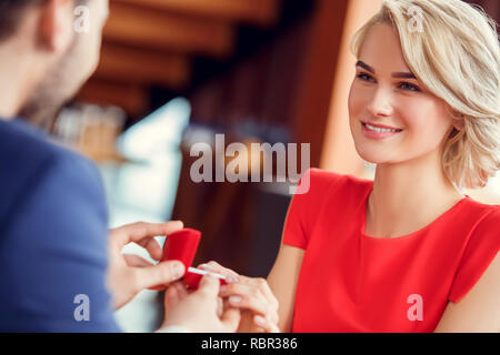 Young man and woman on date in restaurant sitting at table holding box with proposal ring looking at each other smiling happy close-up blurred backgro Stock Photo