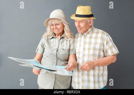 Senior couple tourists wearing beach hats studio standing isolated on gray wall looking at map choosing destination together smiling joyful Stock Photo