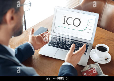 Mature man trader sitting at table at cafe with cup of coffee and laptop using initial coin offering looking at chart confused back view close-up. Stock Photo