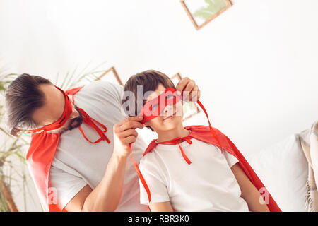 Father and little son wearing superheroe costumes together at home standing man wearing mask on boy's face concentrated close-up Stock Photo
