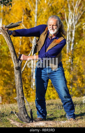 Strong, vital elderly man dressed in hippie style in outdoor exercise using big, heavy branches, on mountain meadow in front of autumn birch forest Stock Photo