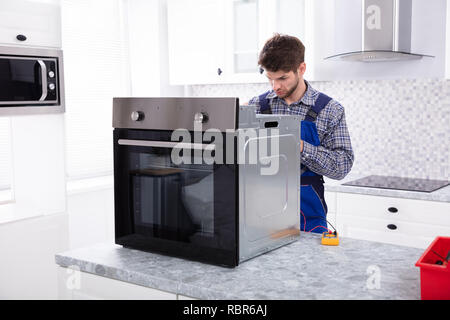 Young Male Repairman Repairing Oven Using Digital Multimeter In Kitchen Stock Photo