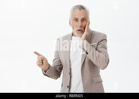 Waist-up shot of amused enthusiastic and happy good-looking fancy old man with beard and grey hair pressing hand to cheek from surprise and amazement pointing left and smiling feeling astonished Stock Photo
