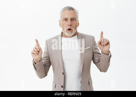 Waist-up shot of stunned emotive shocked granddad in suit over t-shirt with grey beard and short hairstyle open mouth gasping shocked pointing up at curious amazing copy space over white wall Stock Photo