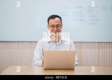 A young Chinese Asian business man is working on his laptop computer in a meeting room in his office. He is smiling as he looks up from his work. Stock Photo