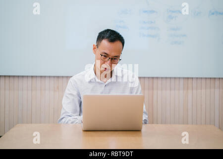 Portrait of an Asian Chinese professional man in shirt and pants sitting and working on his laptop in a meeting room in his office or coworking space. Stock Photo