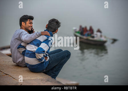 Two friends sitting on the Ghats chatting with each other while speaking on the phone and watching boats passing by on the Ganges in Varanasi, India Stock Photo