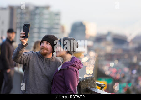 Seattle, Washington, USA. 11th Jan, 2019. Couple takes a selfie among the crowd gathered at the Pike Place MarketFront for the final sunset over the Alaskan Way Viaduct. The highway closed permanently at 10pm on January 11 so crews can move State Route 99 from the viaduct to a state-of-the-art tunnel. The 1950’s-era double-decker freeway was severely damaged in the 2001 Nisqually earthquake and has been maintained long beyond its usable life. A two-mile long, bored road tunnel is replacing the Alaskan Way Viaduct, carrying State Route 99 under downtown Seattle from the SODO neighborhood to Sou Stock Photo
