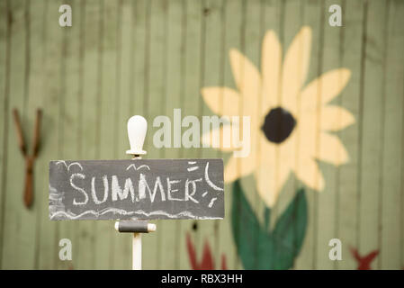 A child like chalk written sign saying Summer, mounted in a backyard garden with colourful painted fence in the blurred background Stock Photo
