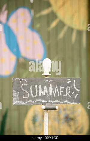 A child like chalk written sign saying Summer, mounted in a backyard garden with colourful painted fence in the blurred background Stock Photo