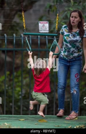 A photo of a strong toddler training on pull up gym rings while his emotional mother is cheering Stock Photo