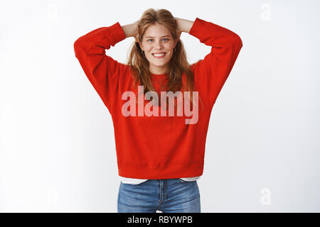 Studio shot of optimistic charming attractive young woman with cute freckles smiling joyfully with nice white smile holding hands on back of head in carefree chill pose wearing oversized red sweater Stock Photo