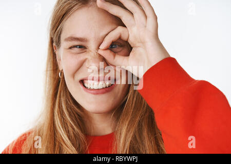 Headshot of happy charismatic and carefree young fair-haired female student with freckles and blue eyes smiling joyfully making okay gesture, looking through it with one eye as peeking over grey wall Stock Photo