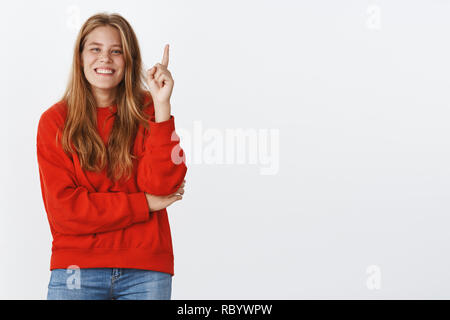 Portrait of carefree charismatic woman rembemer cool joke raising index finger while laughing and smiling adding word with eureka gesture giving suggestion or idea posing in red sweater over grey wall Stock Photo