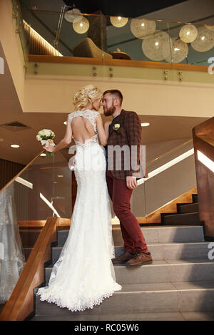 Bride in a chic long dress with a train and the groom stand on big stairs, couple in love hugs on stairs kiss and look at each other. Day of the weddi Stock Photo