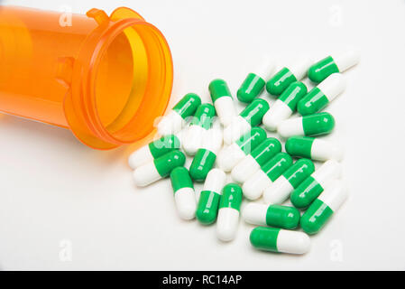 A toppled empty transparent orange medicine bottle spilling out contents of green and white capsules on a plain white background. Stock Photo