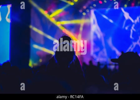 Silhouette of a big crowd at concert against a brightly lit stage. Night time rock concert with people having fun lifting hands up in the air and cheering the musicians Stock Photo