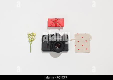 Trendy flat lay composition with retro camera, red gift box, gift canvas bag with red heart shapes and spring field flower on white background. Stock Photo