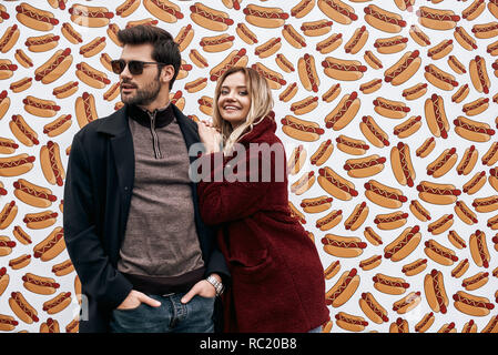 We love hot-doggs. Young couple in eco fur coats at the street brand wall background. Brand wall designed with street food. Cold season. Young man in  Stock Photo