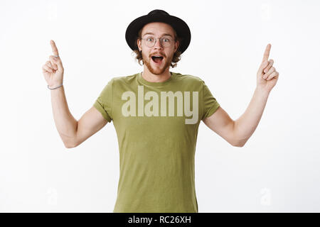 Waist-up shot of surprised and amused handsome charismatic redhead guy with beard in hat and glasses open mouth from amazement raising hands pointing at awesome promotion over gray wall Stock Photo