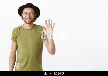 Hi nice to meet you. Portrait of handsome and charismatic young creative male newbie in office waving hand in hello gesture smiling joyfully as greeting new person in club over gray wall Stock Photo