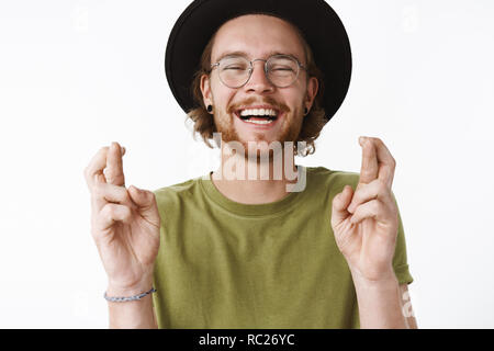 Waist-up shot of optimistic carefree and handsome young bearded guy in glasses and hat laughing out loud with joyful expression as crossing fingers for good luck wishing dream come true over gray wall Stock Photo