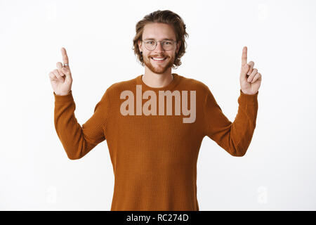 Waist-up shot of charismatic pleased and upbeat handsome young bearded man in glasses with wavy hair, wearing sweater raising hands to point up, making promotion and smiling at camera Stock Photo