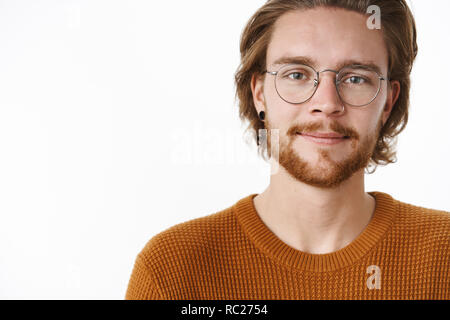 Close-up shot of charming happy and carefree bearded redhead guy with pierced nose in glasses and sweater standing on right side of copy space smiling looking hopefully at camera over gray wall Stock Photo