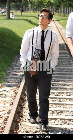 Young man wearing glasses and suspenders walking along a railway track with a vintage camera around his neck Stock Photo