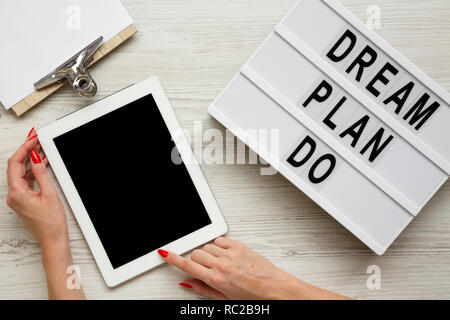 Female hands using tablet with blank screen, modern board with text 'Dream plan do', clipboard over white wooden background, overhead view. From above Stock Photo