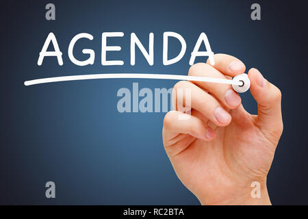Hand writing the word Agenda with white marker on transparent wipe board over dark blue background. Stock Photo