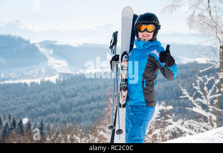 Happy woman skier wearing blue ski suit, black helmet and mask smiling, showing thumbs up in the mountains Stock Photo