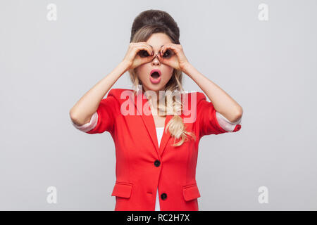 Portrait of surprised beautiful business lady with hairstyle and makeup in red fancy blazer, standing and looking in binoculars gesture. indoor studio Stock Photo