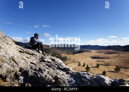 Side view of a woman wearing beanie and a backpack sitting on rocks and enjoying the view over grass covered hills, Slovenia, Milanja. Stock Photo