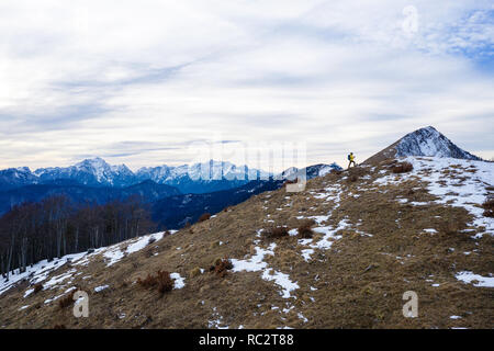 Aerial view of woman hiking to top of grass covered mountain raising hands in winter with great view over the Julian Alps, Slovenia, Pticji vrh Stock Photo