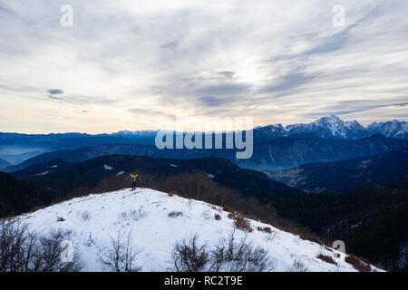 Woman on top of grass covered mountain raising hands in winter with great view over the Julian Alps, Slovenia, Pticji vrh Stock Photo