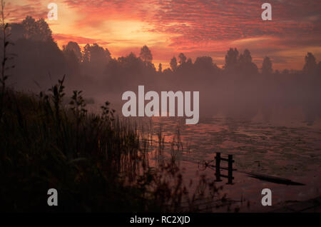 Beautiful orange pink sky over peaceful lake. Foggy autumn sunrise. Trees silhouette, lonely dock quiet forest, swamp plants, smoke mist on still calm water. Russia Moscow area nature landscape Stock Photo