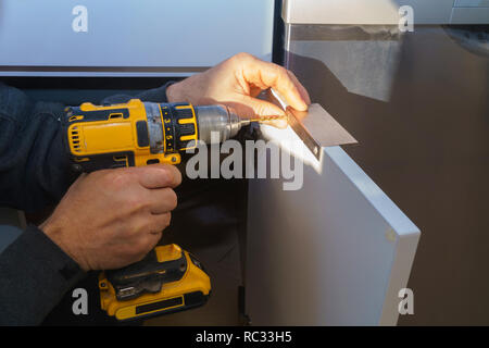 The worker sets a new handle on the white cabinet with a screwdriver installing kitchen cabinets Stock Photo