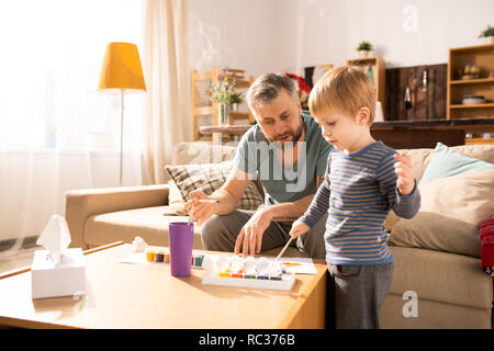 Serious handsome bearded single father sitting on sofa and teaching son to paint using acryl paints, curious boy paining picture with paintbrush Stock Photo