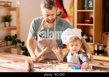 Cute little boy in chefs cap and apron cooking cookies with father and rolling dough in flour, handsome father assisting him Stock Photo