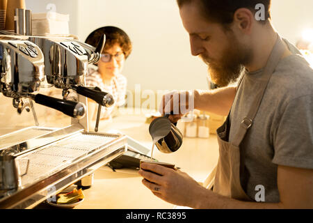 Serious handsome young bearded bartender in apron standing at counter and pouring milk into coffee while preparing coffee for visitor Stock Photo