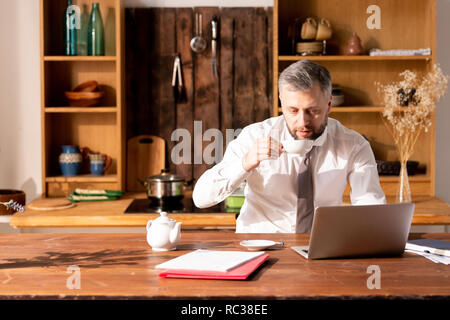 Pensive handsome man using laptop and drinking tea at home Stock Photo