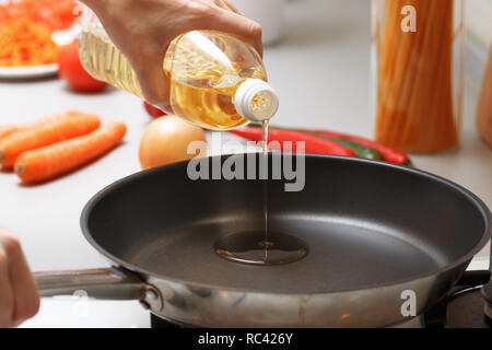 Home cooking. A woman pouring oil from a bottle into the pan in the kitchen, near fresh vegetables and pasta. Stock Photo