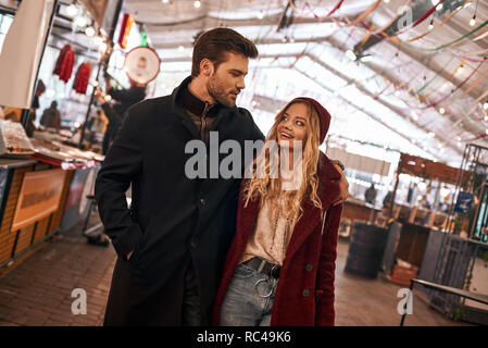 Stroll through the market together. Happy young joyful couple walking around street food market. Autumn season, blond haired woman wearing red cap, he Stock Photo