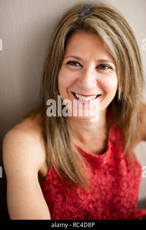 Bust portrait of smiling woman at the age of 50, with blond hair and makeup, wearing red sleeveless dress, leaning back on grey wall, looking away wit Stock Photo