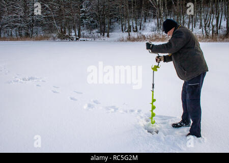 Man bores ice with drill for fishing in winter snowy morning, frozen lake Stock Photo