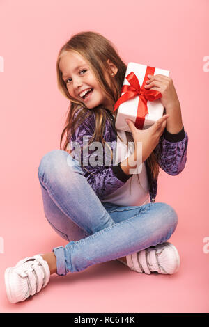 Pretty little girl sitting isolated over pink background, holding gift box Stock Photo