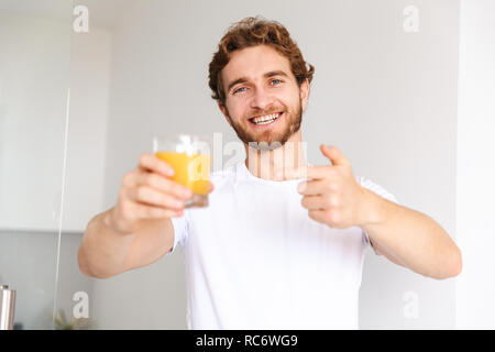 Image of a handsome young bearded man at home drinking juice showing thumbs up. Stock Photo
